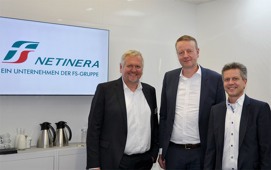 Gruppenbild vor einem weißen Hintergrund. Links befindet sich ein Bildschirm mit dem Logo von NETINERA, darunter eine kleine Anrichte mit Gläsern, Tassen und Kaffeekannen.