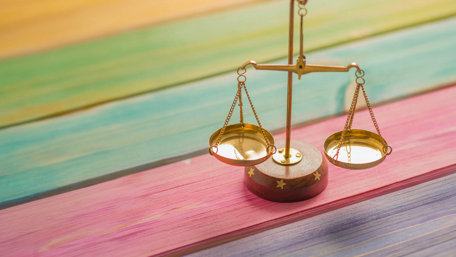 Une petite balance dorée sur une table en bois dont les lattes sont peintes aux couleurs de l'arc-en-ciel et représentent l'égalité des chances