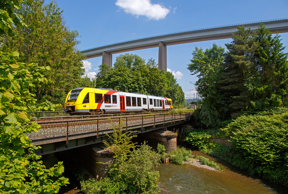 Un train HLB blanc et jaune passe sur un pont qui enjambe une rivière dans une forêt.