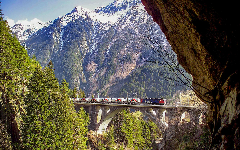 Eine Eisenbahn überquert eine Brücke von links nach rechts vor einem Bergpanorama.