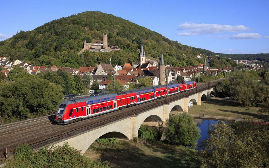 Ein Roter DB Regio Twindexx Doppelstock-Zug überquert eine Brücke vor einer malerischen Landschaft mit Burgruine im Hintergrund.
