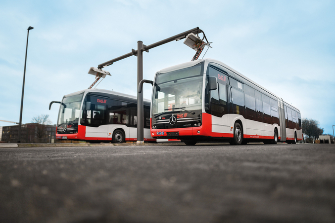 Deux bus électriques rouges et blancs d'EvoBus sont chargés sur une borne de recharge dans un dépôt.