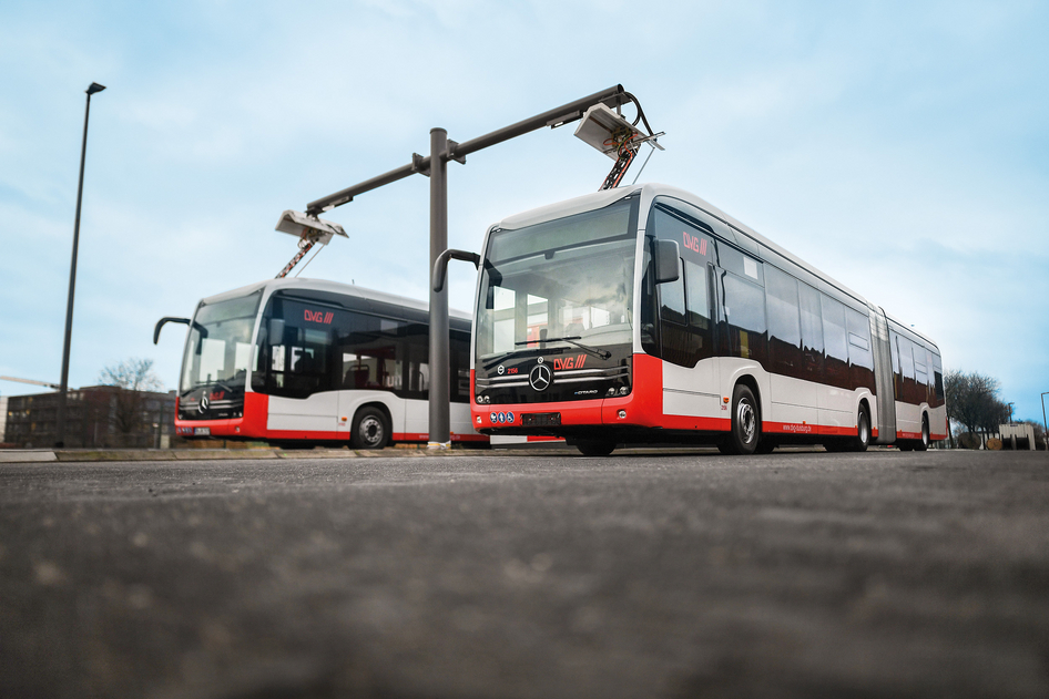 Deux bus électriques rouges et blancs d'EvoBus sont chargés sur une borne de recharge dans un dépôt.