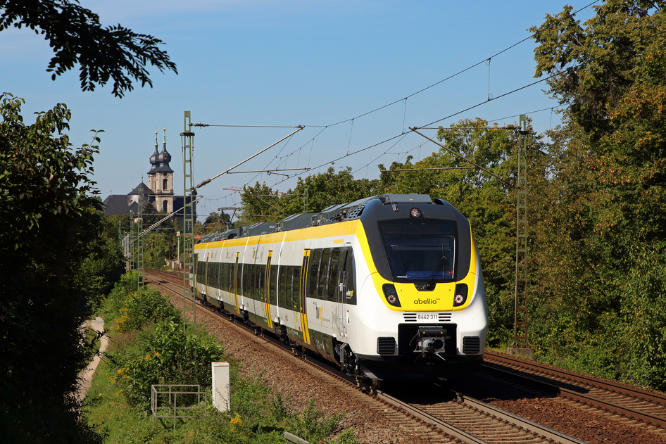 Un train blanc et jaune de la SWEG circule entre les arbres avec une petite gare en arrière-plan.
