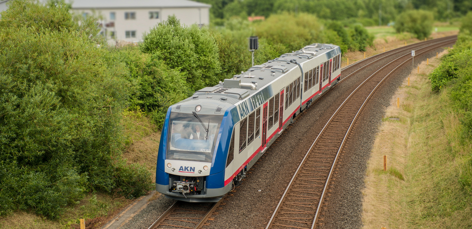 Ein blau-weiß-roter AKN Zug fährt nahe einer Ortschaft im Grünen.
