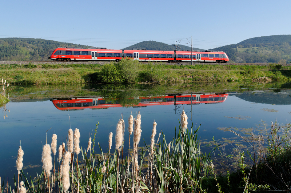 Un train régional DB rouge passe devant une rivière et se reflète dans celle-ci.