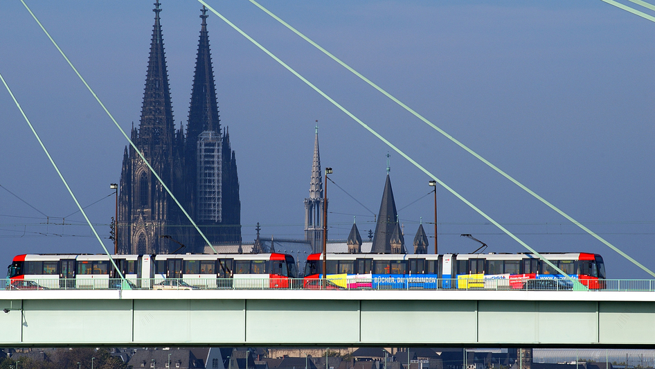 Die Kölner Straßenbahn fährt auf einer Brücke mit dem Kölner Dom im Hintergrund.