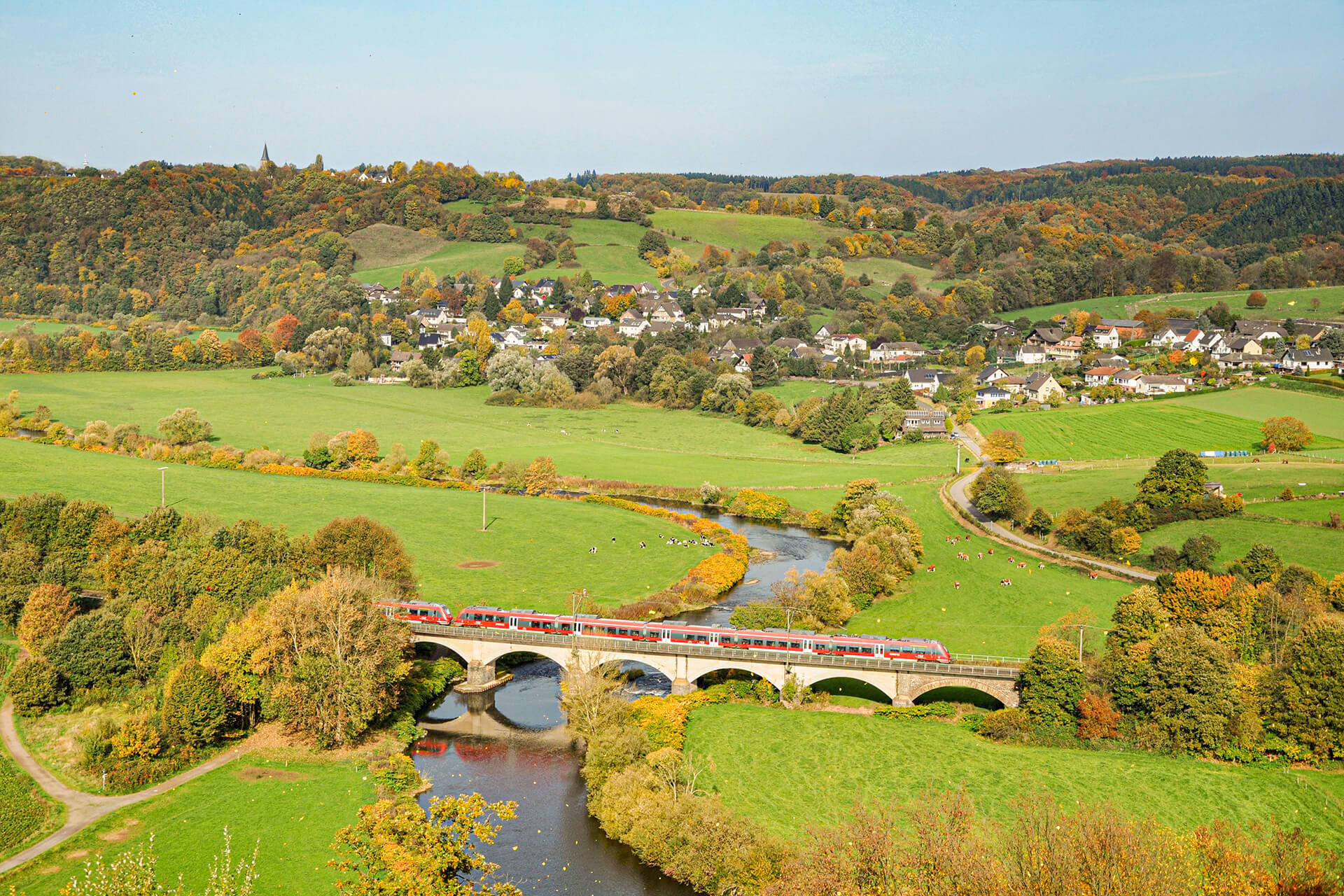 Train rouge DB Regio traversant un pont sur une rivière dans la campagne avec une petite ville en arrière-plan